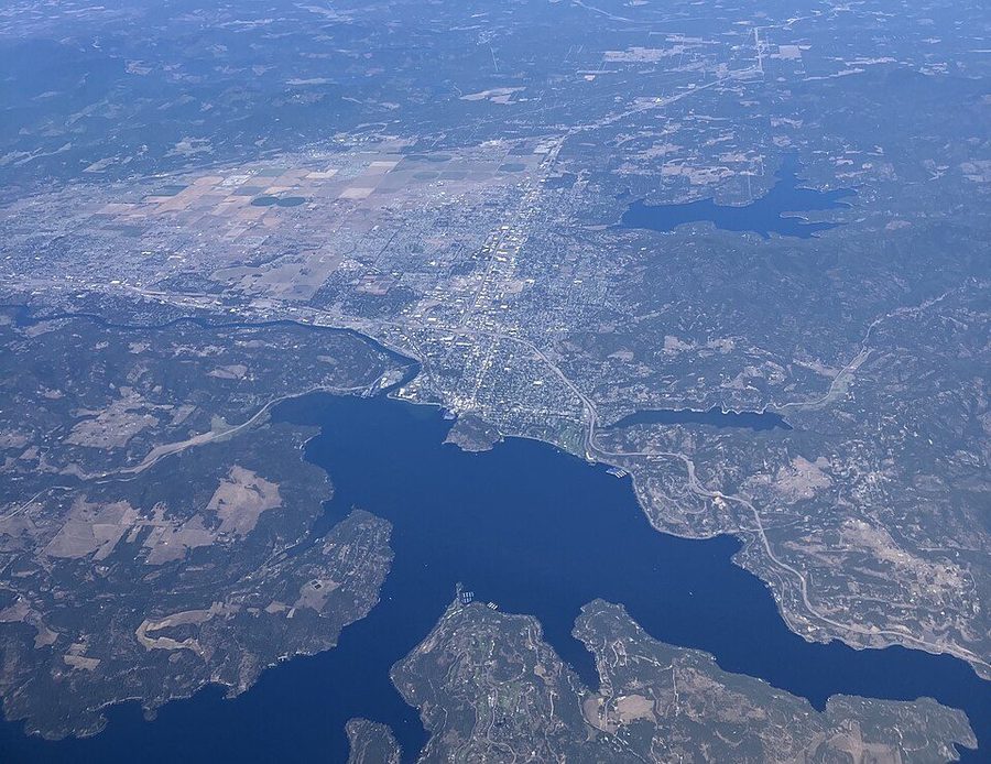 Aerial view of Coeur d'Alene, Idaho — the town on the shore of a large blue lake surrounded by forested hills.