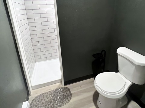 Inside the bathhouse at Round Mountain Retreat — a white subway-tiled walk-in shower with a pebble bath mat and a flush toilet against deep charcoal-green walls on pale wood-look floors.