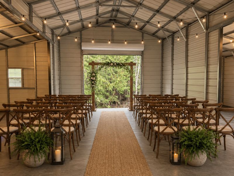 Inside the covered pavilion set for a wedding ceremony — two blocks of wooden cross-back chairs either side of a jute aisle runner, lanterns and potted ferns at the front, and a floral arch in the open doorway looking out to green forest.