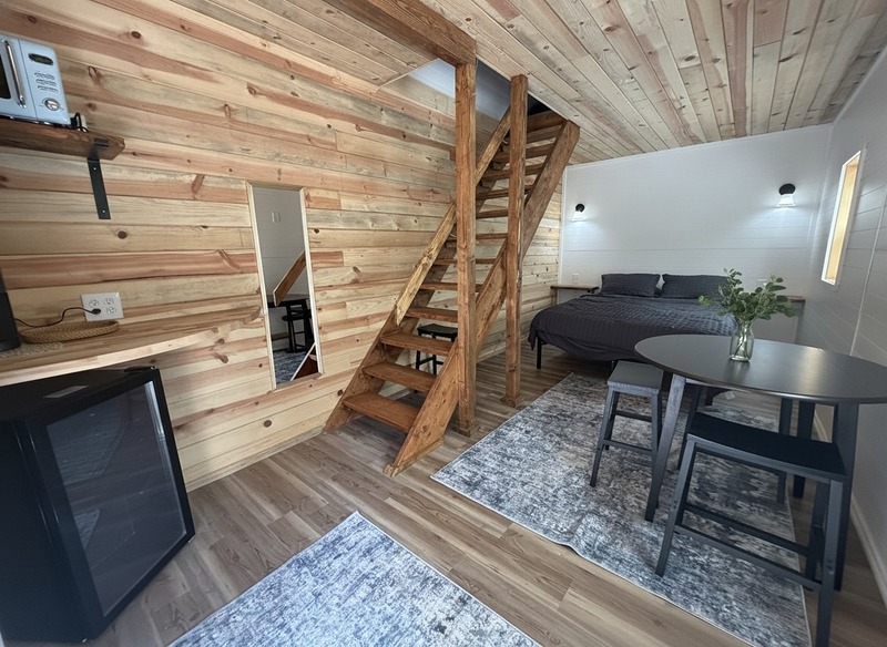 Inside The Caribou at Round Mountain Retreat — a cedar-lined two-storey cabin with a timber loft staircase, queen bed, round dining table and a small drinks fridge.