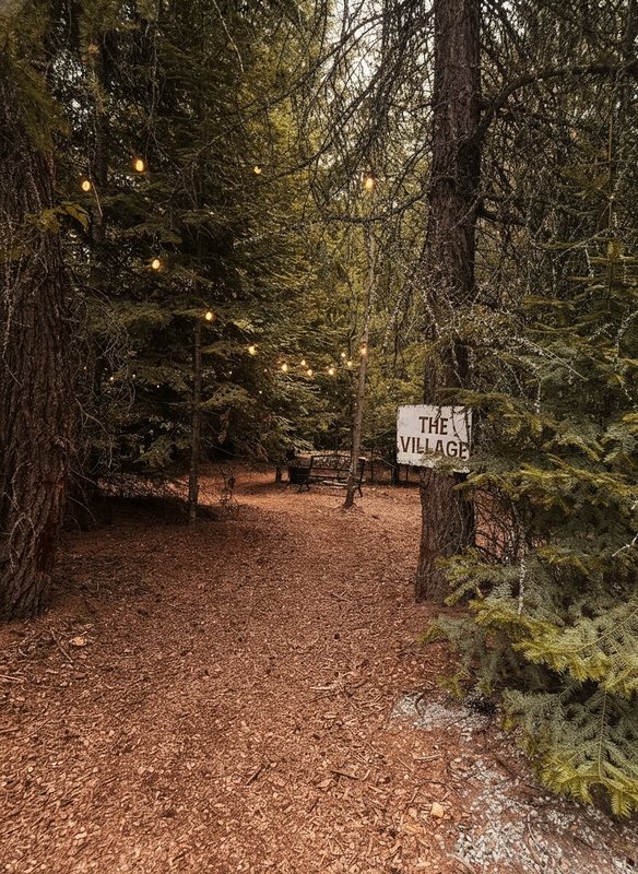 The soft woodchip path leading into The Village at Round Mountain Retreat, warm string lights glowing through the pines above a hand-painted sign reading THE VILLAGE.