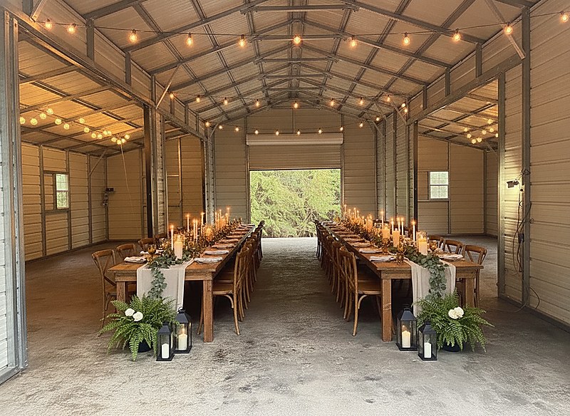 The pavilion set for a wedding dinner — two long timber banquet tables with linen runners, eucalyptus, taper candles and lanterns, the big end door rolled up on to green forest and warm string lights overhead.