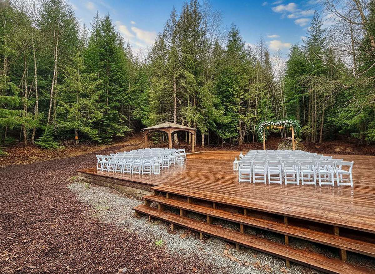 The open-air ceremony deck at Round Mountain Retreat set with white chairs either side of the aisle, a flower-dressed arbour at the treeline and evergreen forest all around.