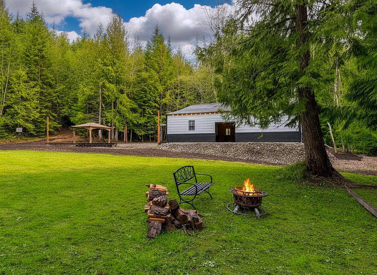 The event building at Round Mountain Retreat on a summer afternoon — a white building at the top of a green lawn, string lights along the eaves, a fire bowl and bench on the grass and dense evergreen forest behind.