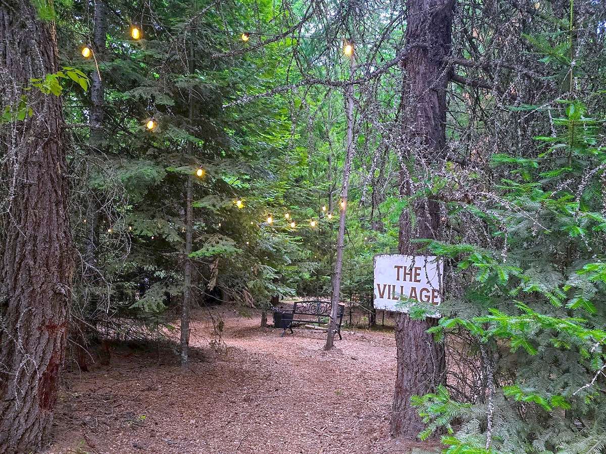The woodchip path into The Village at Round Mountain Retreat, string lights glowing through the pines above a hand-painted sign reading THE VILLAGE — the clearing the cabins and luxury yurts stand in.