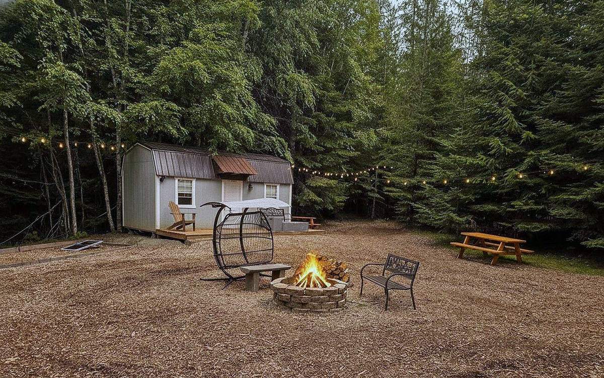 A fire burning in the big stone fire ring in the village at Round Mountain Retreat, chairs pulled up around it and string lights glowing in the pines at dusk.