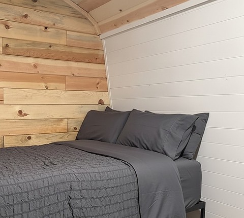 A twin bed in the loft of The Caribou at Round Mountain Retreat, made up in grey linen against a cedar plank wall under the curved white ceiling.