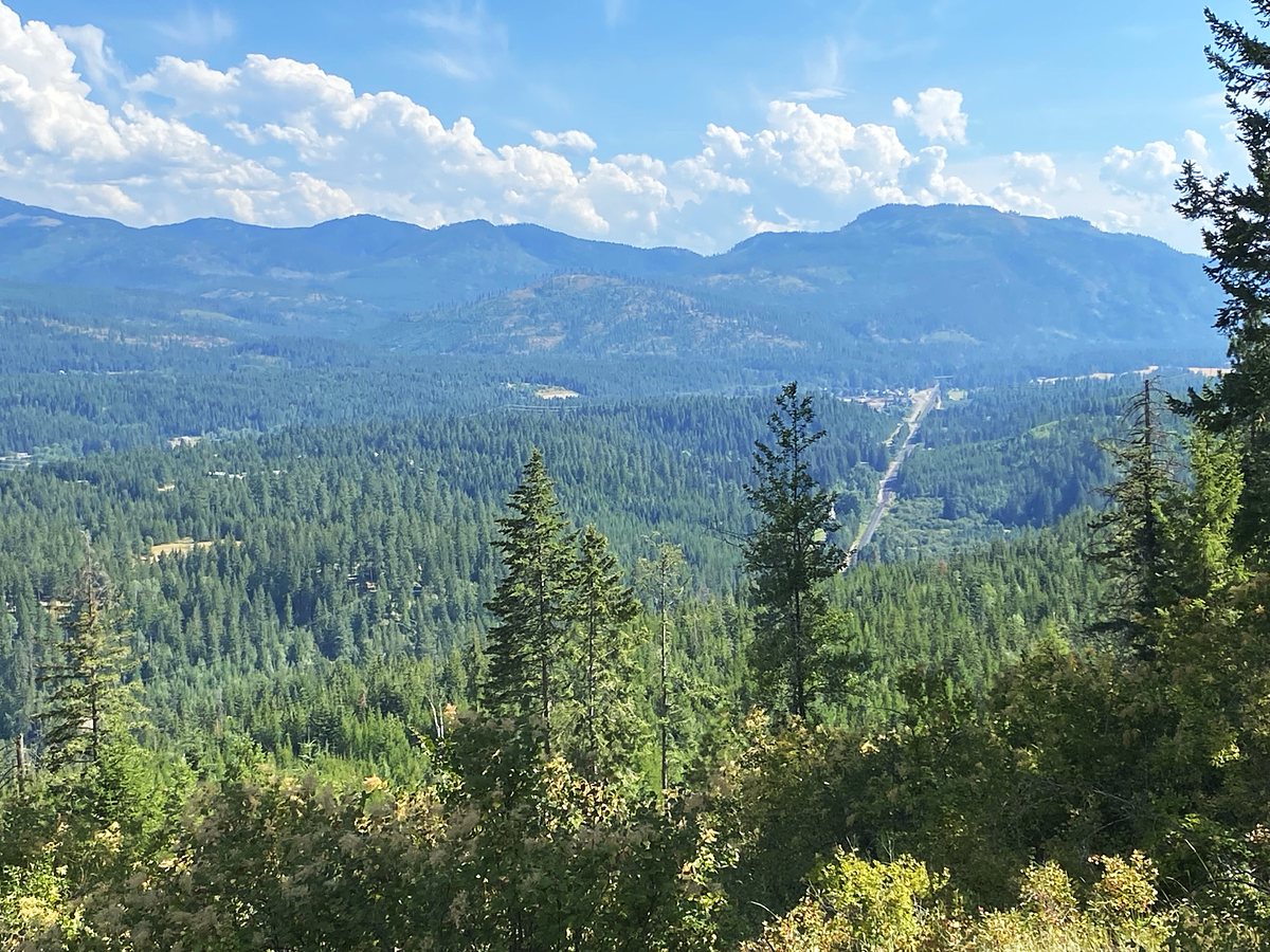 A hundred miles of Idaho Panhandle from the ridge above Round Mountain Retreat — forested mountains layered to the horizon under tall white cumulus