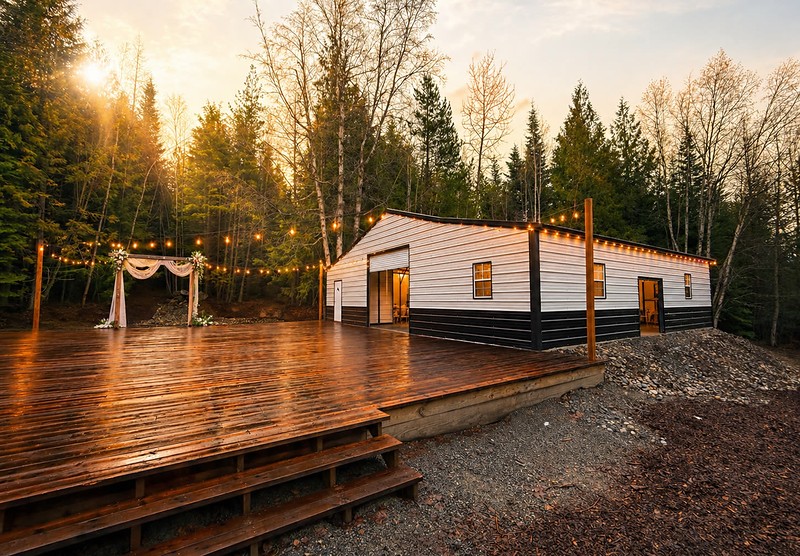 The covered pavilion at Round Mountain Retreat at golden hour — string lights strung across from the ceremony deck to the eaves, warm light spilling from the rolled-up end door, and the sun setting through the evergreens behind.