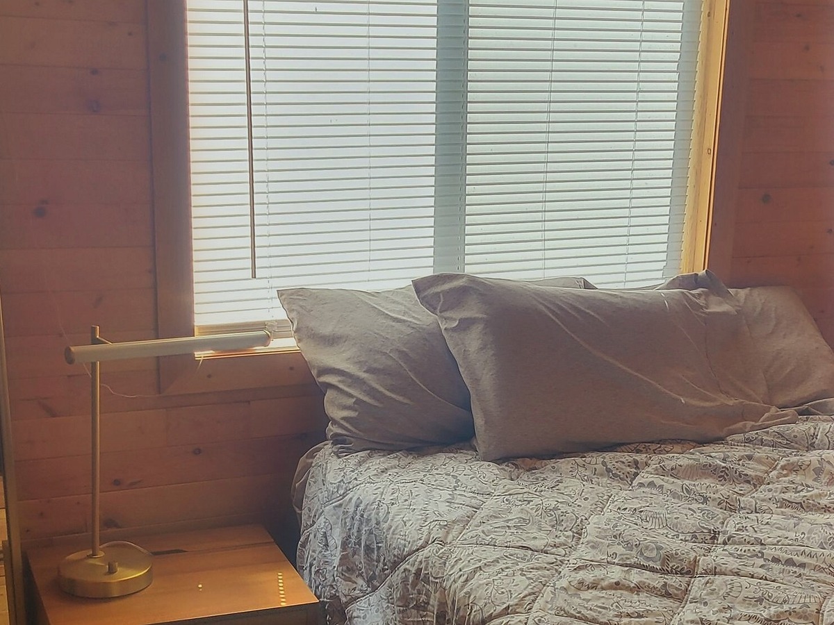 A bedroom in The Elk, made up in grey linen beneath a wide blind-shaded window, with a wooden nightstand and a brass reading lamp