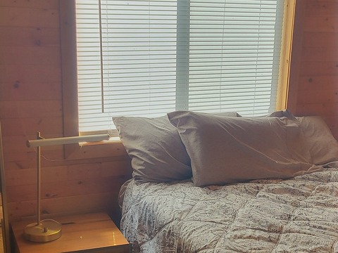 A bedroom in The Elk at Round Mountain Retreat, made up in grey linen beneath a wide blind-shaded window, with a wooden nightstand and a brass reading lamp.