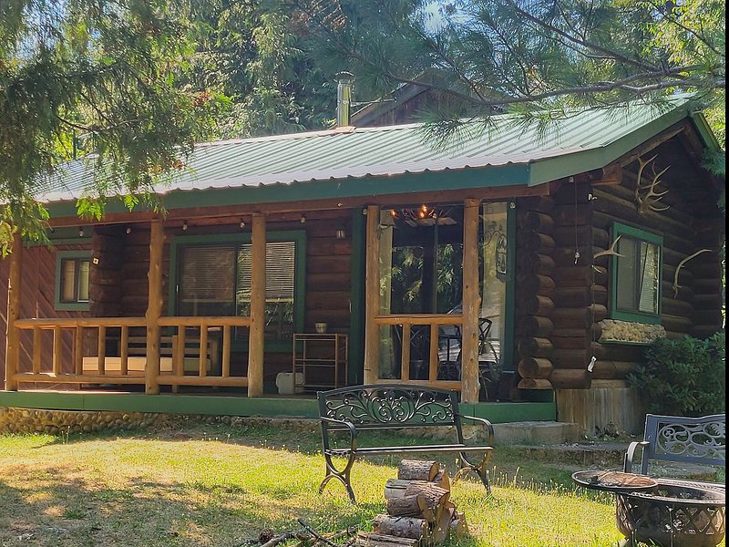 The Elk at Round Mountain Retreat — a hand-built log cabin with a green metal roof, a covered porch on peeled-log posts and a fire pit with benches on the lawn.